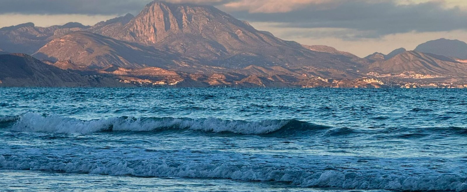 Serene view of Alicante's coastline with calming waves and majestic mountains at sunset.